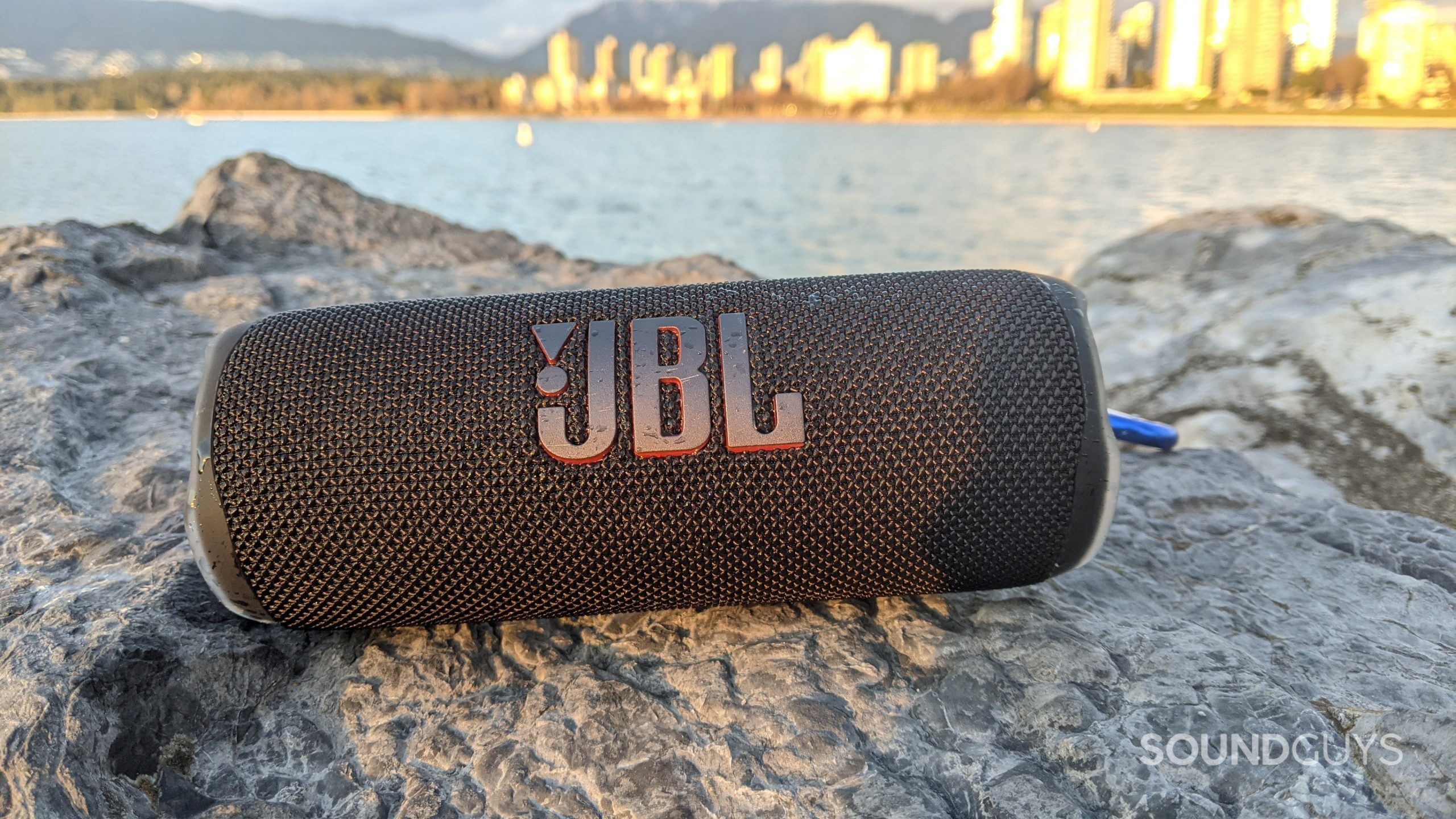 JBL Flip 6 Outdoor On Rocks City Skyline A JBL Flip 6 Bluetooth speaker sitting on a rocky surface with a sunny city skyline viisble in the distance.