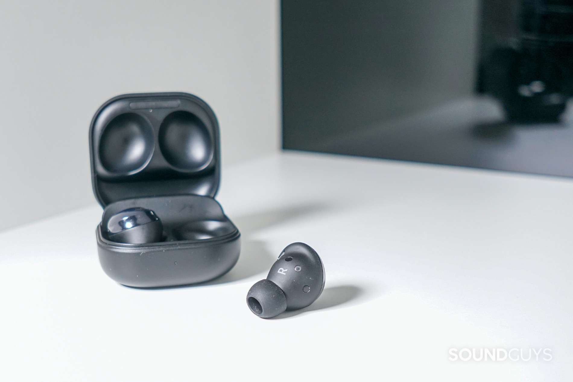 Samsung Galaxy Buds Pro one out of case The Samsung Galaxy Buds Pro true Wireless earbuds sit on a white shelf in front of a reflective black surface, with one earbud in the case.